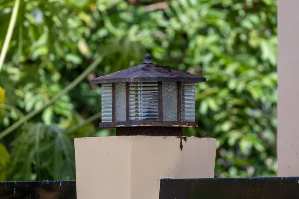 Outdated outdoor lighting system showing a rusted garden lamp with green foliage.